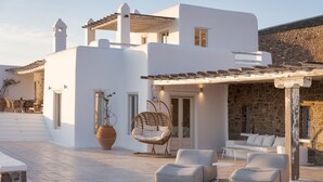 A modern building with a stone facade, a white exterior, and a patio area with seating and a hanging chair. - Mykonian Icon Villa (Mykonos)