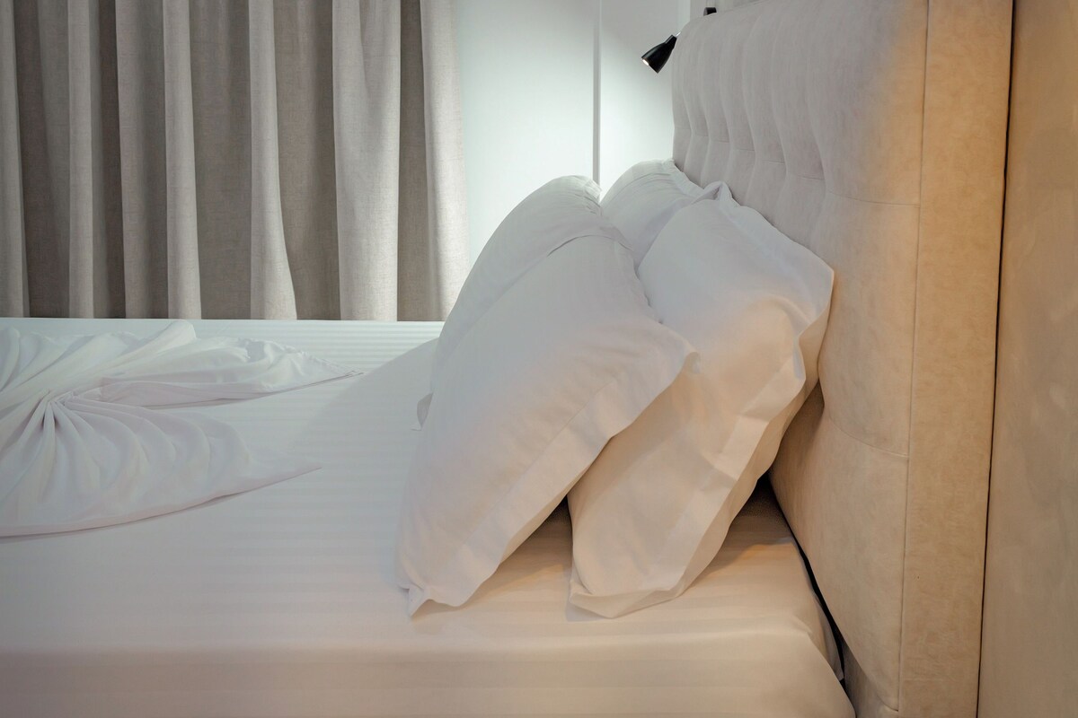 A neatly made bed with white linens, a wooden nightstand with a remote control, and a wall-mounted light fixture.