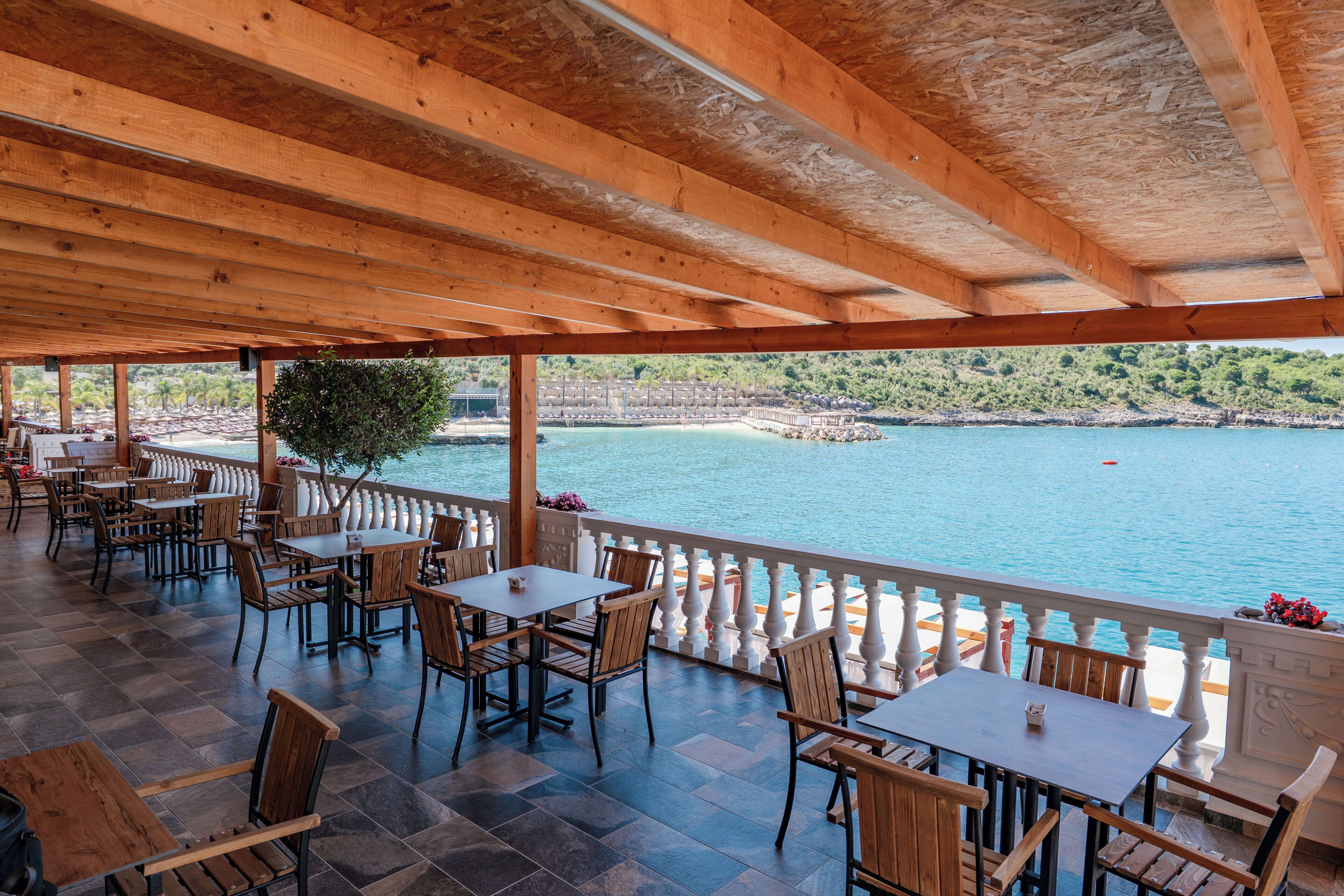 A covered outdoor seating area with tables and chairs, overlooking a body of water and a rocky shoreline.