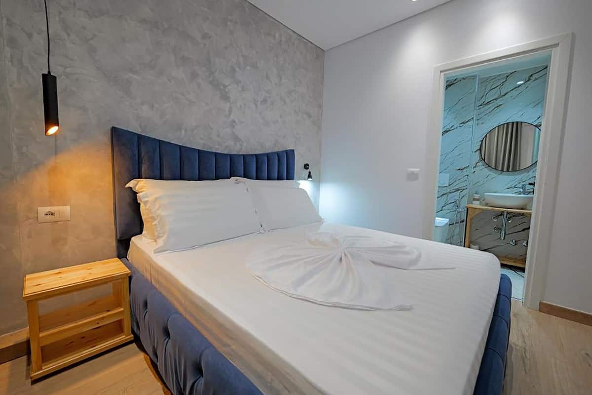 Un dormitorio moderno con una cama grande, una mesita de noche de madera, una pared de mármol y un baño con lavamanos de mármol.