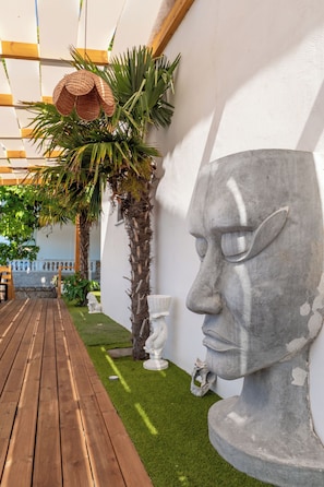 Une terrasse en bois, un mur blanc orné d’une sculpture en pierre et un palmier.