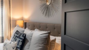 Une chambre à coucher avec une tête de lit capitonnée, un miroir orné d’un motif en forme de soleil, et un lit agrémenté de plusieurs coussins.
