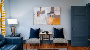 A living room with a blue couch, two patterned chairs, a small table with a lamp, and a large abstract painting on the wall.
