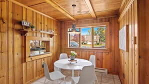 Interior de madera con una mesa redonda para comer, sillas blancas, una lámpara colgante y una ventana con vistas a casas.