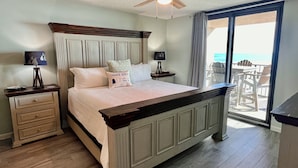 A bedroom with a bed, a nightstand, a dresser, a ceiling fan, and a view of the beach through a large window.
