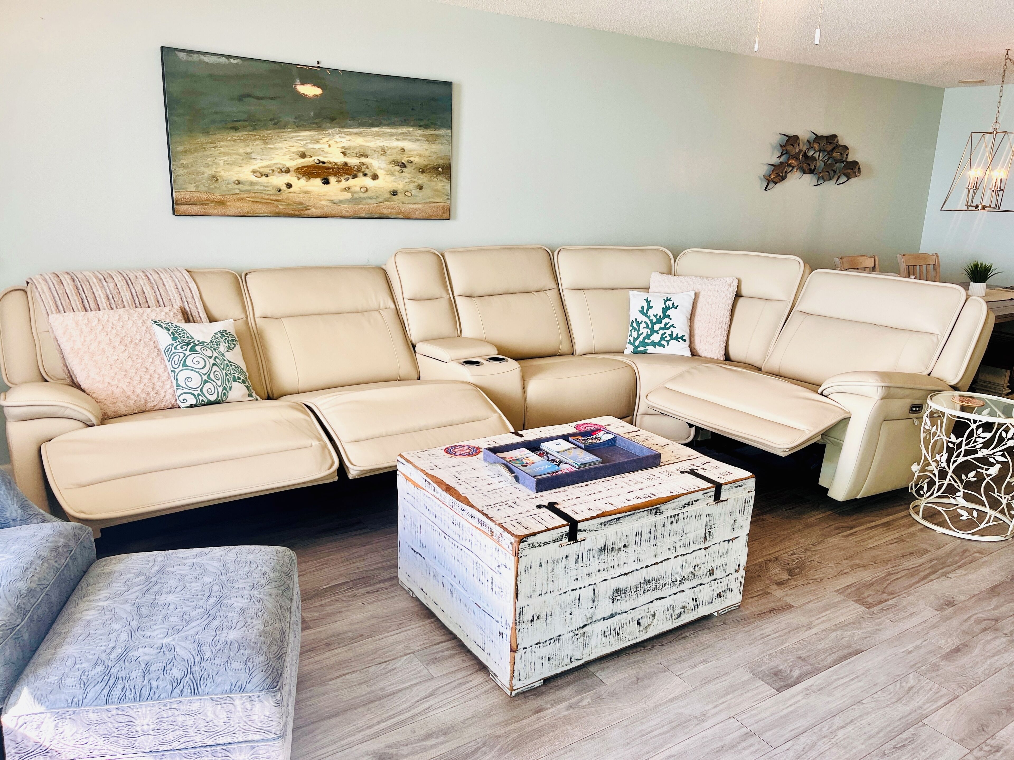Un soggiorno con un divano modulare beige, una poltrona fantasia, un settimino bianco e un quadro raffigurante una spiaggia.
