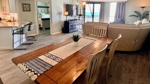 A spacious dining area with a wooden table, chairs, and a chandelier. A sofa and a plant are visible in the background.