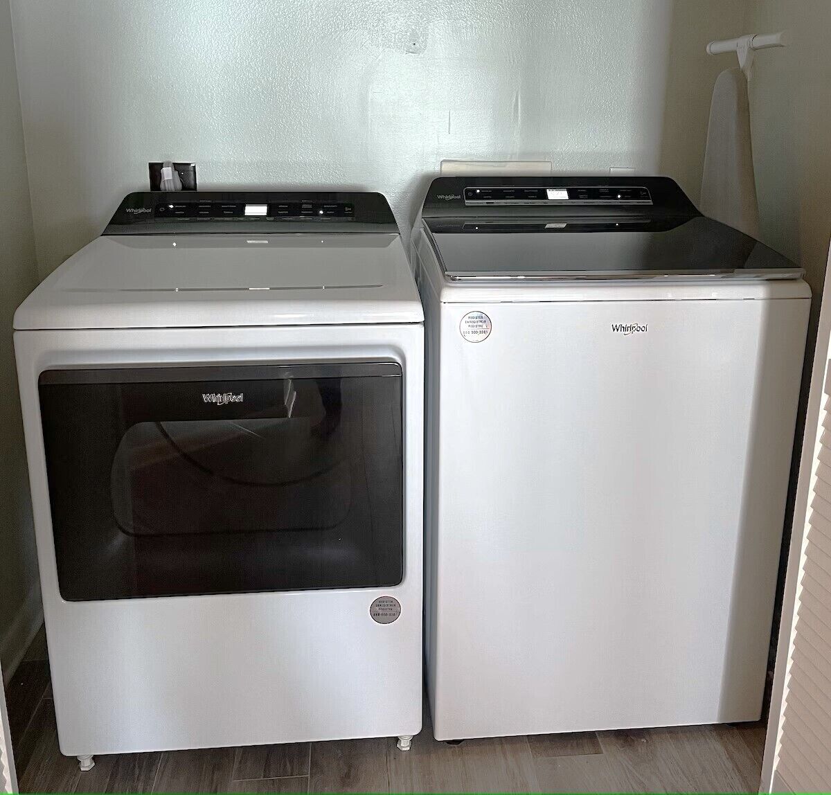 Two Whirlpool washing machines in a laundry room with a wooden floor and a towel rack in the background.