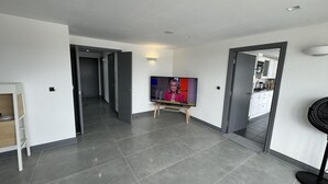 A modern apartment with a flat-screen TV on a stand, grey tiled floors, and a kitchen area visible through an open door. - Luxurious 3 Bedroom Penthouse in City Centre (Chelmsford)