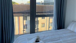 A hotel room with a large bed, a window with curtains, and a view of buildings outside. - Luxurious 3 Bedroom Penthouse in City Centre (Chelmsford)