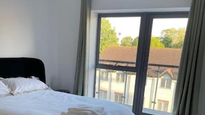 A bedroom with a large bed, grey carpet, and a view of buildings through a window. - Luxurious 3 Bedroom Penthouse in City Centre (Chelmsford)