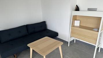 Quarto moderno com um sofá azul escuro, uma pequena mesa de madeira e uma estante branca com um laptop e uma garrafa.