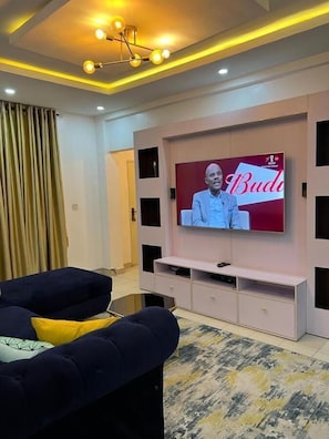 A living room with a flat-screen TV mounted on a wall, a sofa, and a patterned rug. - Amazing luxury 2 bedroom apartment VI (Lagos)