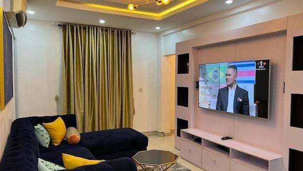 A modern living room with a dark sofa, a glass coffee table, and a large flat-screen TV mounted on a pink entertainment unit.