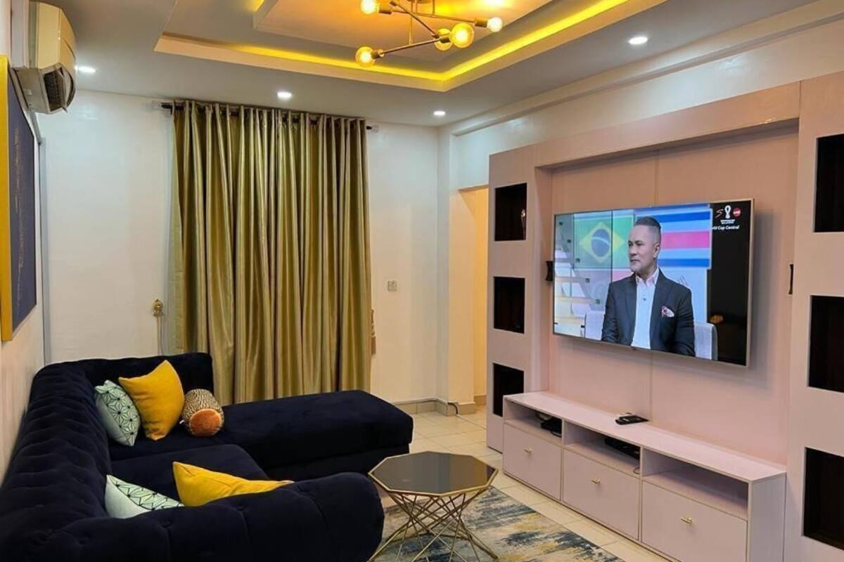 A modern living room with a dark sofa, a glass coffee table, and a large flat-screen TV mounted on a pink entertainment unit.