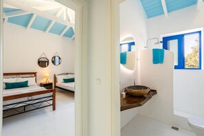 Un dormitorio con cama de madera, lámpara de noche y espejo. Junto a él, un ba ño con lavamanos de madera, espejo y toallas azules.