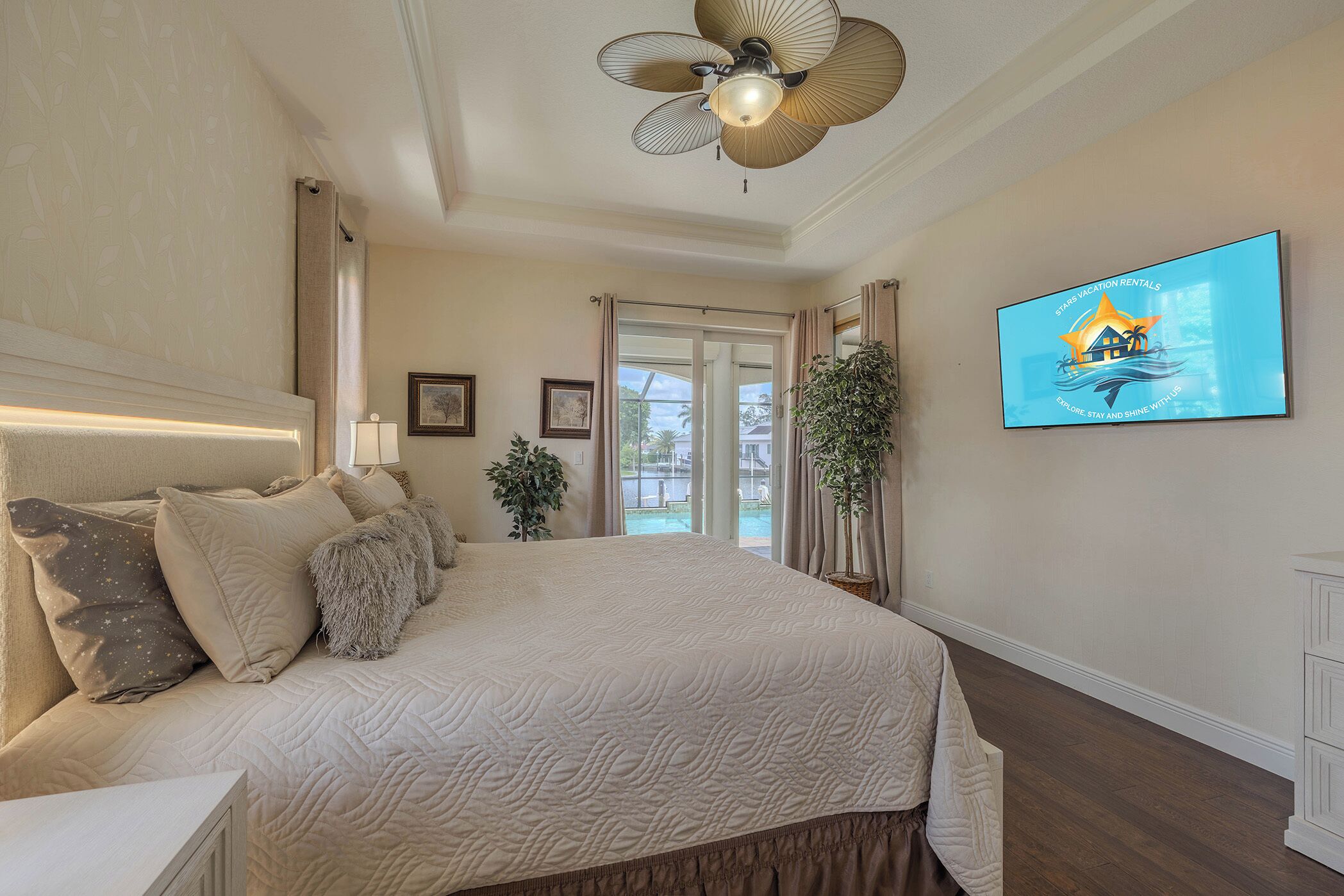 A bedroom with a large bed, a ceiling fan, a TV on the wall, and a view of the outdoors.