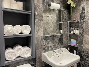 Ein Badezimmer mit Marmorwänden, einem weißen Waschbecken und Regalen mit Handtüchern und Pflegeprodukten.