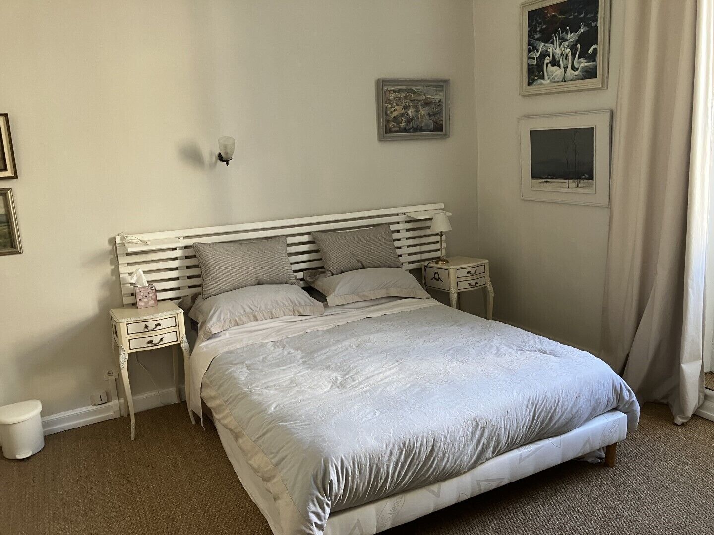 A bedroom with a white bed, two bedside tables, a nightstand, a wall-mounted lamp, and two framed pictures.