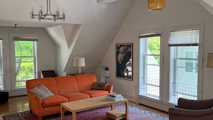 A spacious living room with a high ceiling, a large orange sofa, a wooden coffee table, and a dining area with a wooden table and chairs.