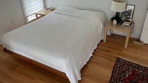 A neatly made bed in a room with wooden flooring, a bedside table with a lamp and books, and a window with blinds.