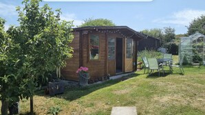 Une cabane en bois avec une porte en verre, un petit jardin avec une table et des chaises, et une serre en arrière-plan.