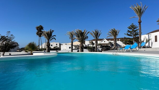 A clear blue pool surrounded by palm trees and white buildings with a clear sky above.