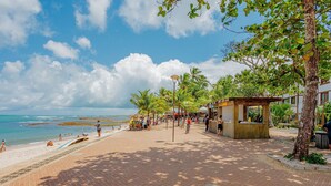 A beachside walkway with palm trees, a beach, and a building with a covered seating area. - PF55 Ótima Casa 4 Suítes -Praia do Forte (Mata de São João)