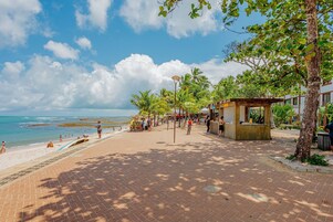 Un paseo marítimo frente a la playa, con palmeras, arena y un edificio que cuenta con un porche con asientos.
