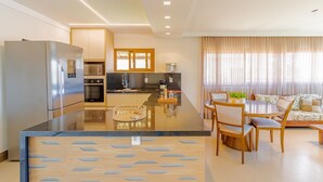 A modern kitchen with a central island, stainless steel appliances, and a dining area with a round table and chairs. - PF55 Ótima Casa 4 Suítes -Praia do Forte (Mata de São João)