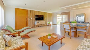 A spacious living area with a wooden sofa, a dining table with chairs, a TV mounted on the wall, and a kitchen area visible in the background. - PF55 Ótima Casa 4 Suítes -Praia do Forte (Mata de São João)