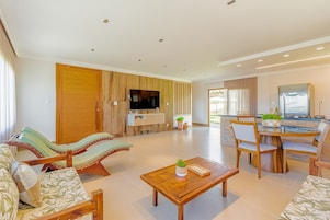 Amplia sala de estar con un sofá de madera, una mesa de comedor con sillas, un televisor montado en la pared y una cocina visible al fondo.
