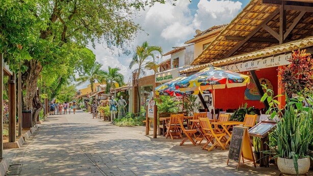A street lined with outdoor seating, a restaurant named "RESTAURANTE," and a sign for "ENERGIA NATURAL." - PF55 Ótima Casa 4 Suítes -Praia do Forte (Mata de São João)