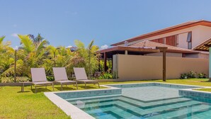 A swimming pool with a tiled deck, surrounded by grass and palm trees, and a modern building with a sloped roof in the background. - PF55 Ótima Casa 4 Suítes -Praia do Forte (Mata de São João)
