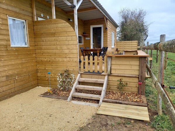A wooden cabin with a porch, a small outdoor seating area, and a dirt ground.
