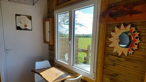 A room with a wooden floor, a table with two chairs, a window with a view of a green field, and a wall decoration. - Chalet en Ferme Équestre au Plus Proche des Chevaux (Yèvres)