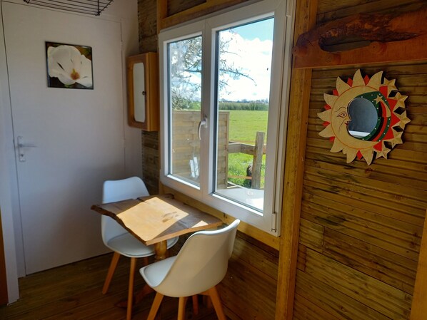 Una habitación con piso de madera, una mesa con dos sillas, un ventanal con vista a un campo verde y un adorno en la pared.