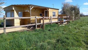 A wooden cabin with a fenced porch, a car parked outside, and a grassy field in the background. - Chalet en Ferme Équestre au Plus Proche des Chevaux (Yèvres)