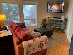 Un salon chaleureux avec une cheminée, un téléviseur fixé au mur, un canapé avec des coussins, un petit sapin de Noël et un meuble avec des portes en verre.