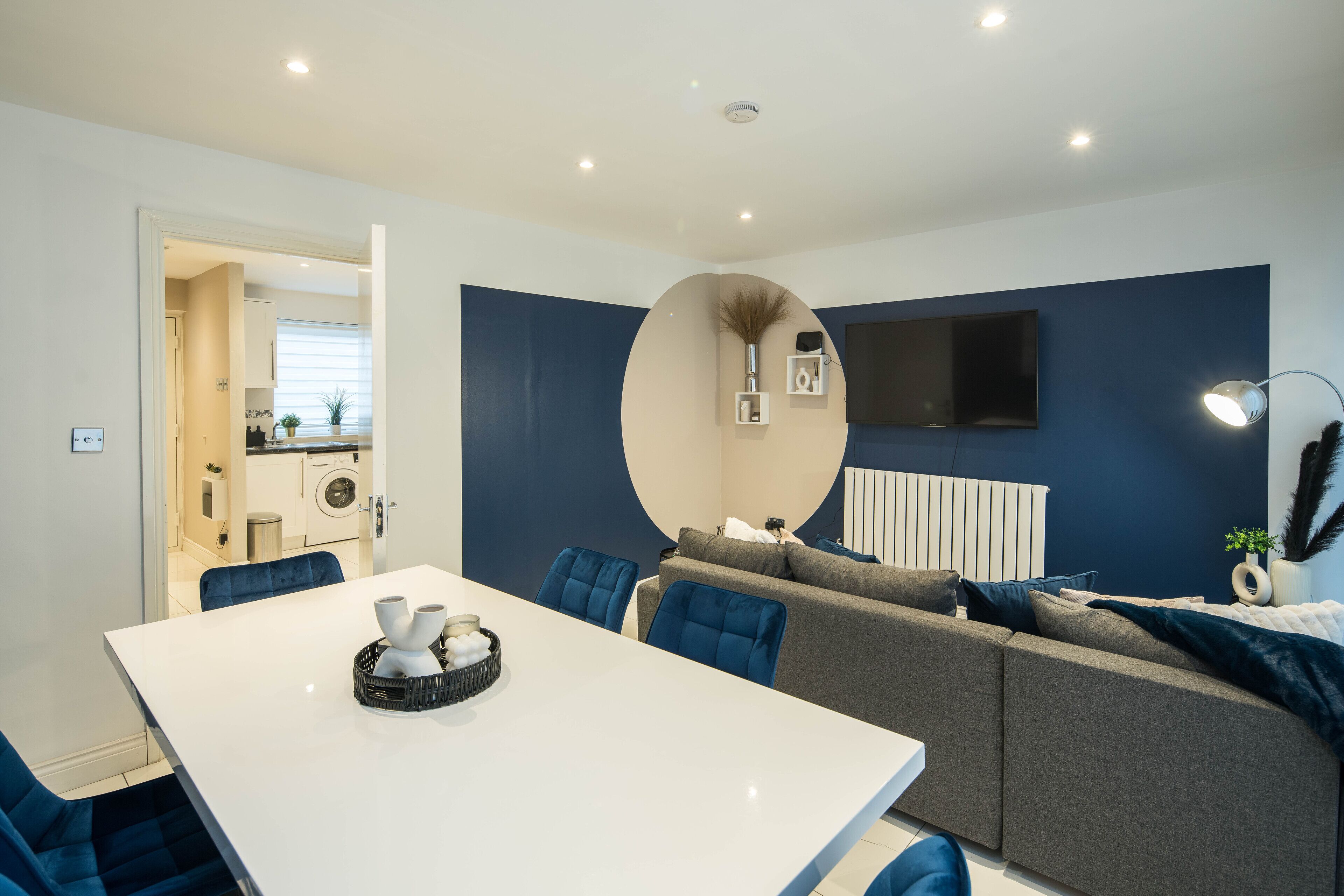 A modern living room with a white dining table, blue chairs, and a grey sofa.