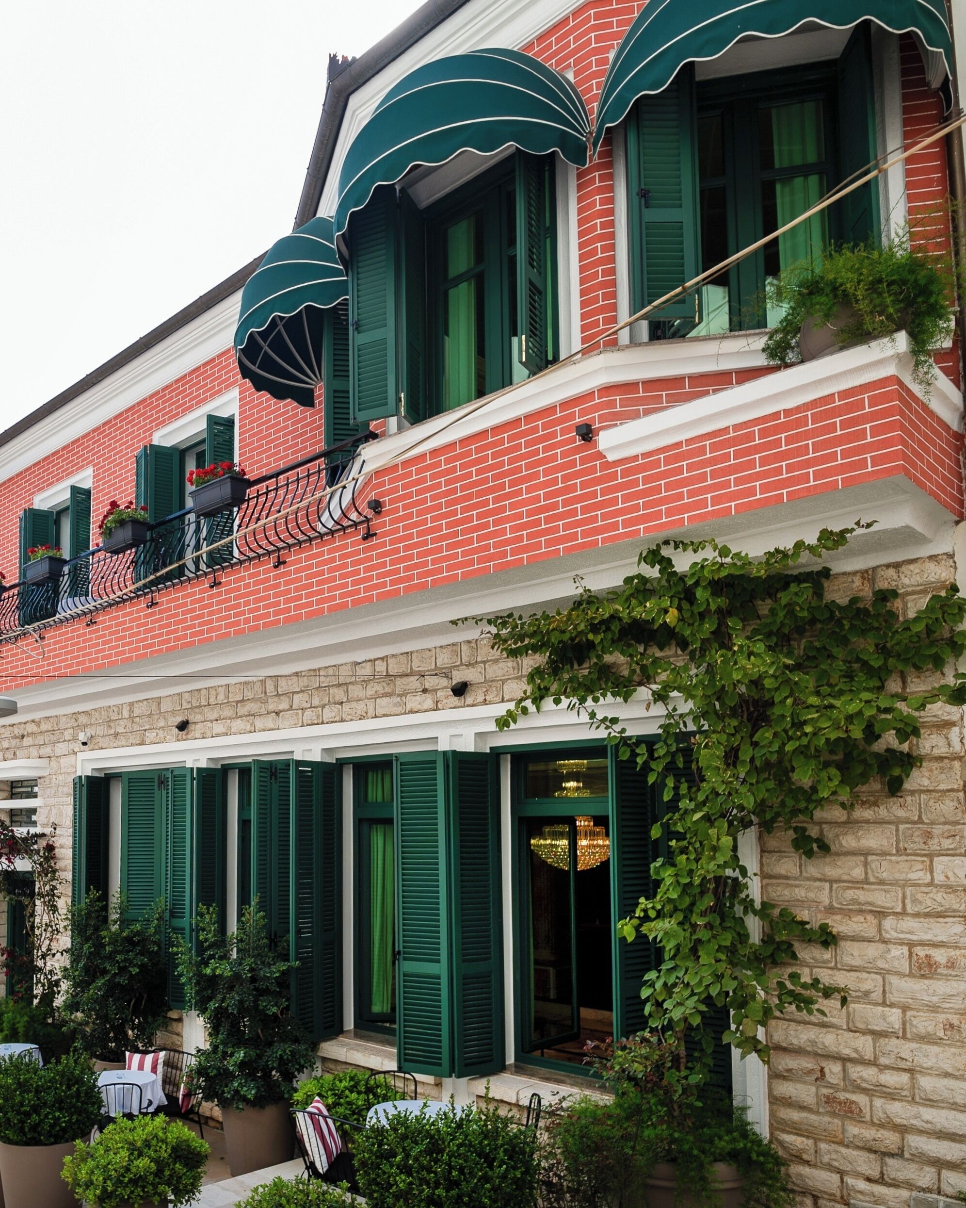A two-story building with red brick walls, green shutters, and a balcony with outdoor seating.