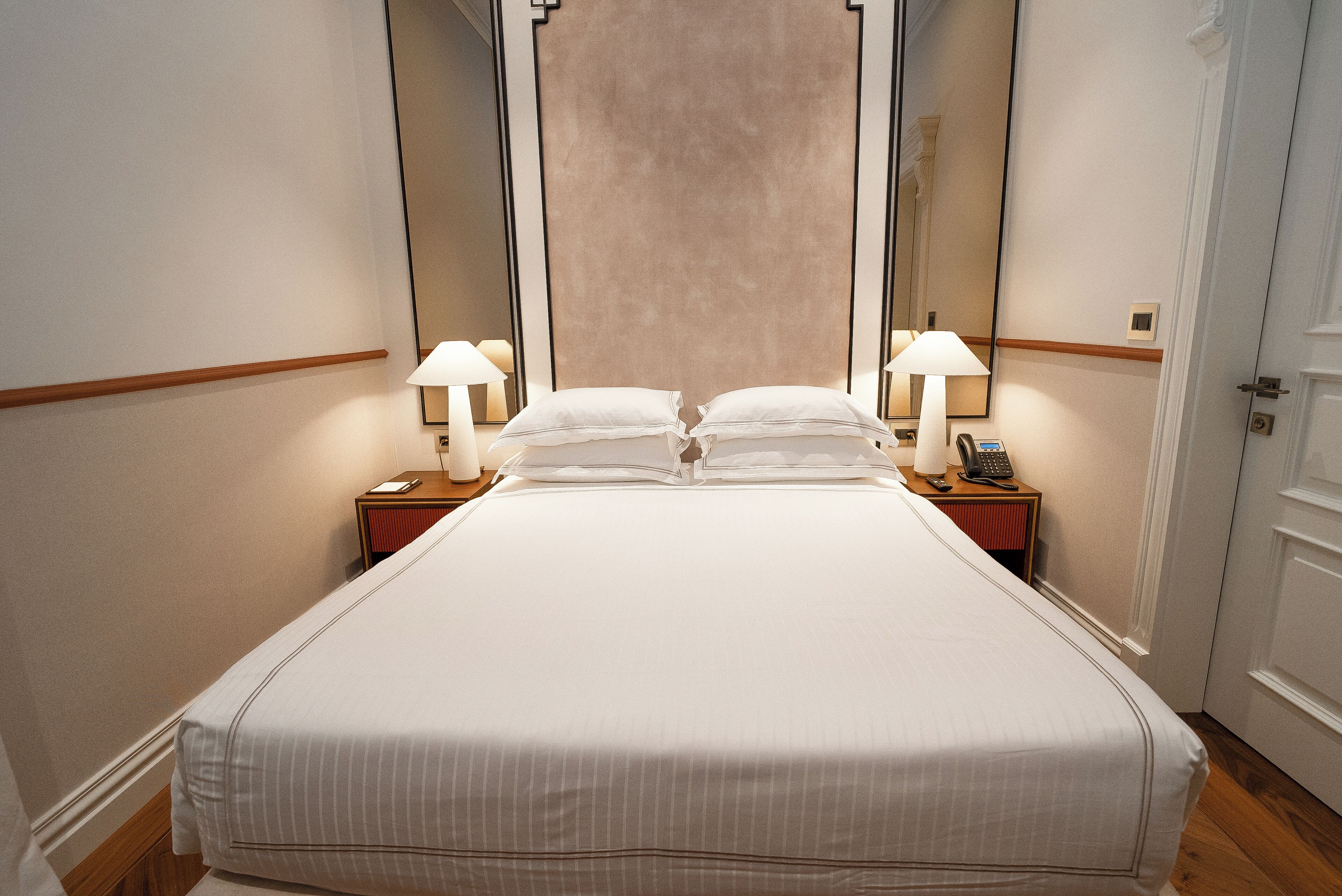 A neatly made bed with white linens, two bedside lamps, a nightstand with a telephone, and a large mirror on the wall.