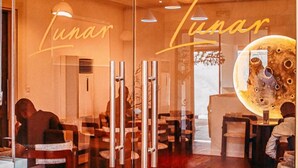 L’intérieur d’un restaurant présente un mur de verre sur lequel est inscrit le nom « Lunar Lunar ». À l’intérieur, on trouve des tables et des chaises disposées pour accueillir les clients.