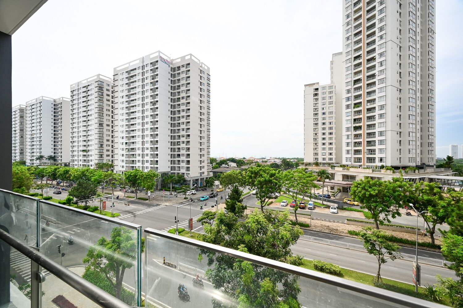 A high-rise residential building complex with multiple apartment buildings, a well-maintained road with vehicles, and lush greenery.