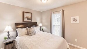 Un dormitorio con una cama grande, dos mesitas de noche con lámparas, una lámpara de techo, una ventana con cortinas y un cuadro en la pared.