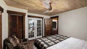 A bedroom with a wooden ceiling, a bed with a patterned blanket, a chair, and a door leading to a balcony.