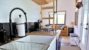 Een moderne keuken met een grote witte spoelbak, een zwarte kraan en houten aanrechtbladen. Er is een raam met uitzicht naar buiten, en een zithoek met kussens en een tafel.