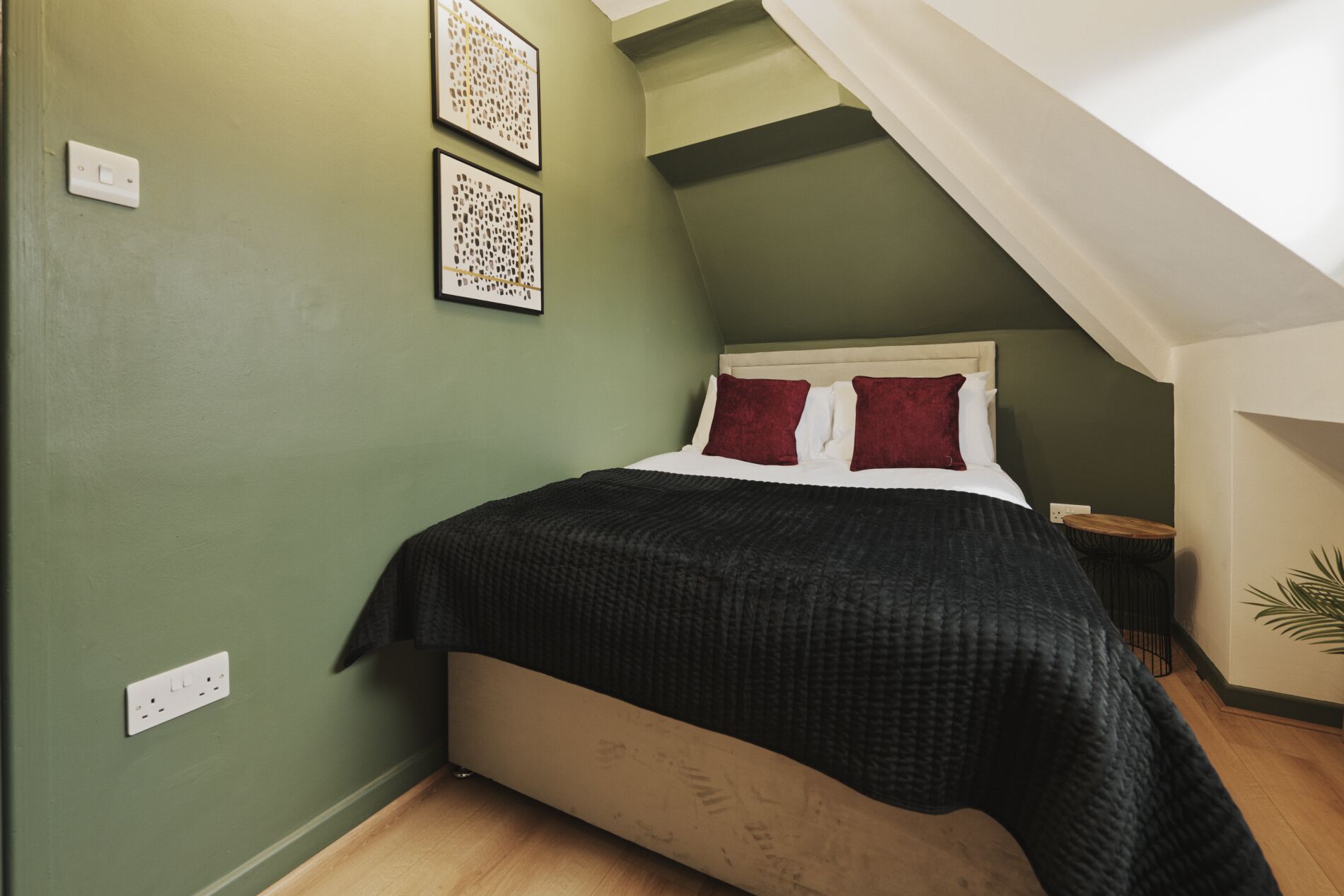 A bedroom with a sloped ceiling, a bed with a white headboard and red pillows, a dark blanket, and two framed artworks on the wall.