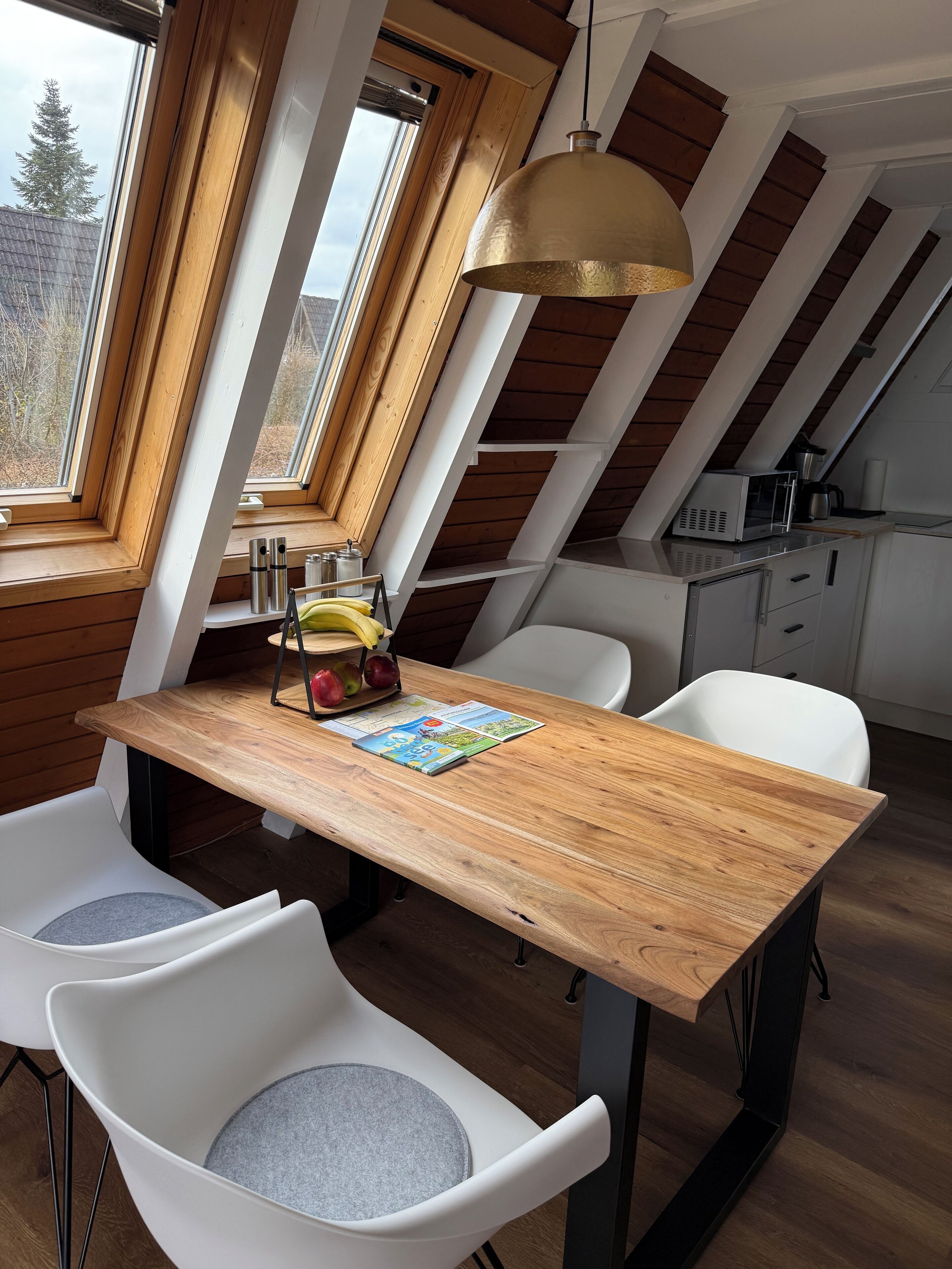 A dining area with a wooden table, two white chairs with grey cushions, a hanging pendant light, and a view of the outdoors through large windows.