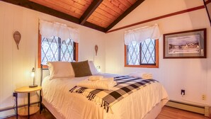 Un dormitorio acogedor con una cama grande, dos ventanas con cortinas, una mesita de noche con una lámpara y un cuadro enmarcado en la pared.